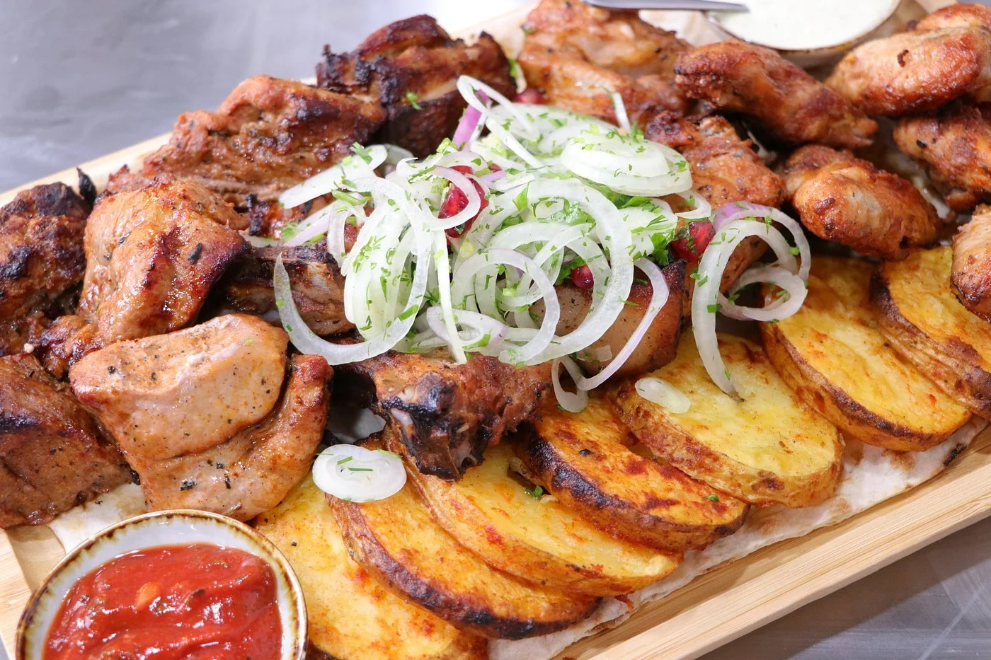 A platter of grilled chicken and steak with roasted potato slices topped with sliced onions and green herbs, served with a small bowl of red dipping sauce.