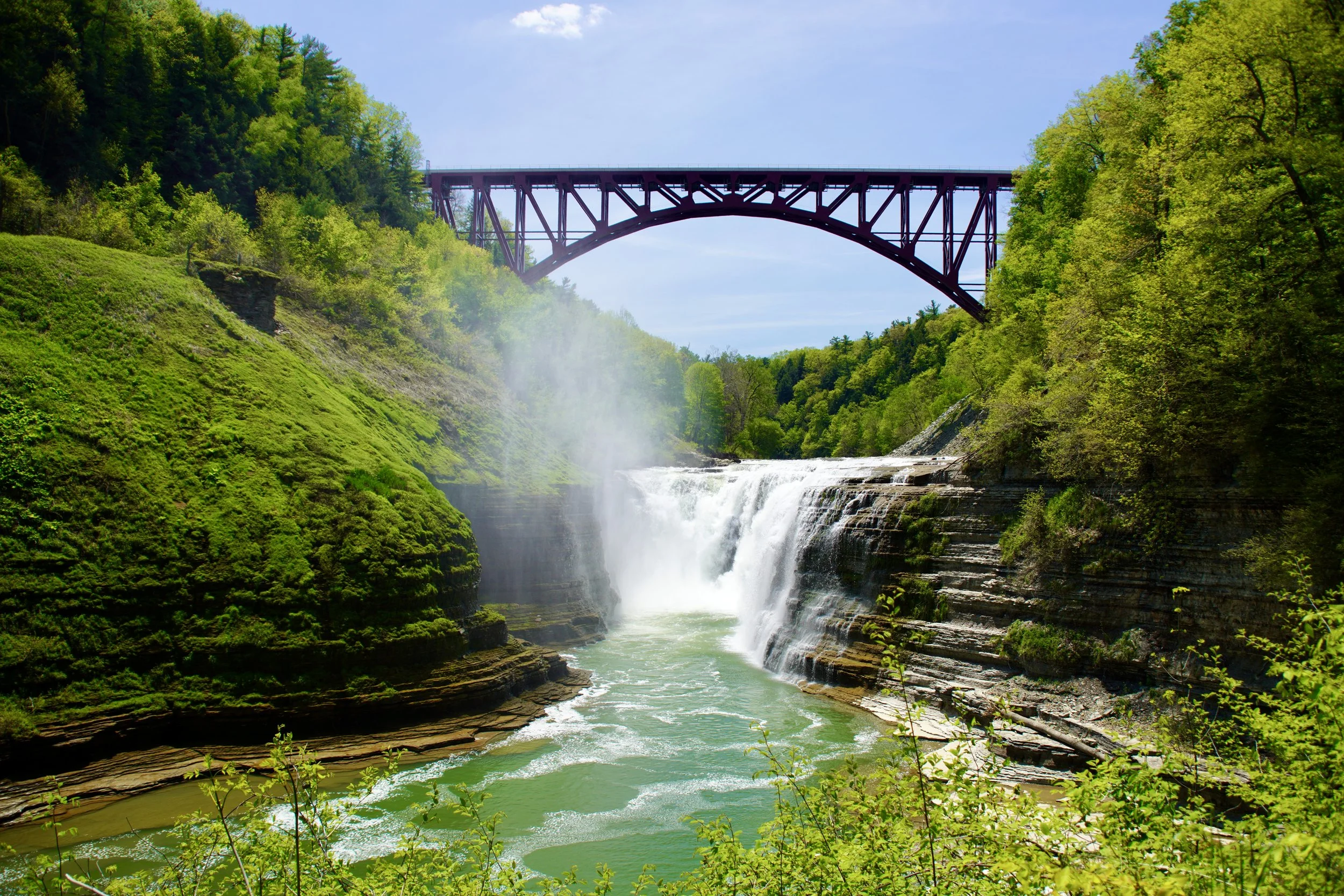 Waterfall in a lush green valley with a bridge overhead and a partly cloudy blue sky.