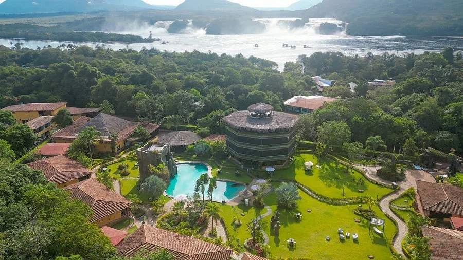 Aerial view of a tropical resort with a large swimming pool, surrounded by lush green gardens and small buildings with red-tiled roofs, and a river with waterfalls in the background.