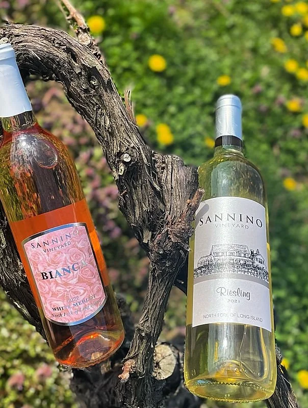Two bottles of wine hanging from a gnarled tree branch in a garden with yellow and pink flowers in the background.