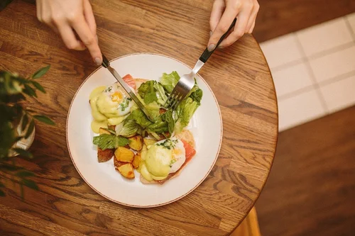 Overhead view of a person cutting into a dish with poached eggs, roasted potatoes, and salad on a white plate on a wooden table.