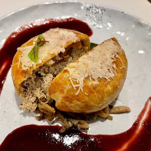 A breaded food item, possibly a stuffed pastry or bread bowl, filled with shredded meat and mushrooms, topped with grated cheese and garnished with microgreens, served with a drizzle of red sauce on a white plate.