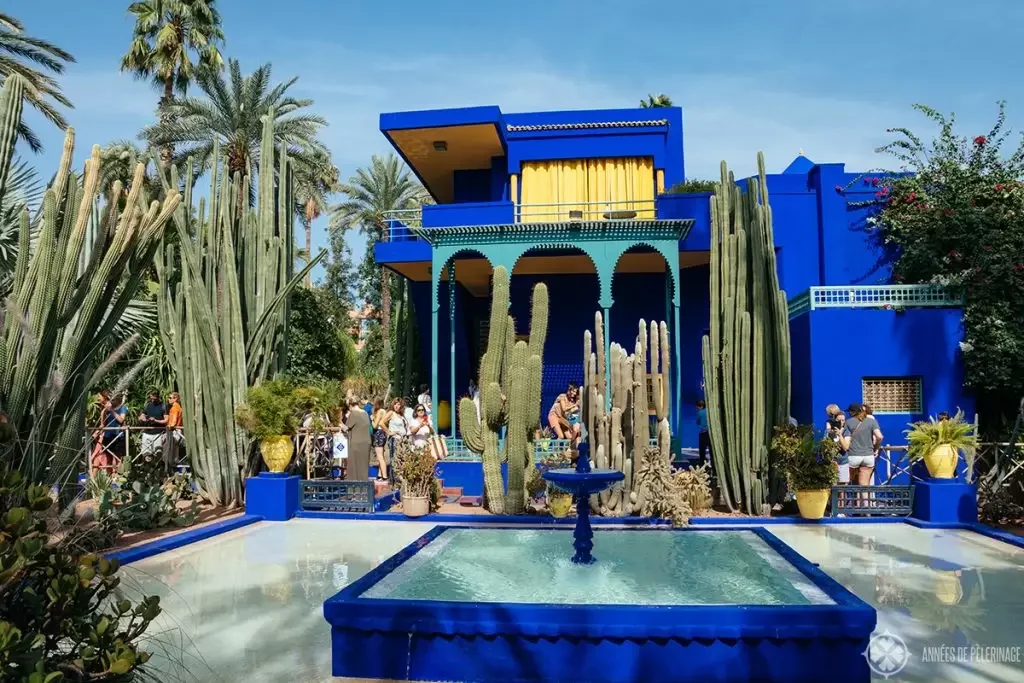 A vibrant building painted in bright blue with a yellow curtain, surrounded by tall cactus plants and palm trees. There is a small water fountain in the foreground, and people are walking and gathering around the area on a sunny day.
