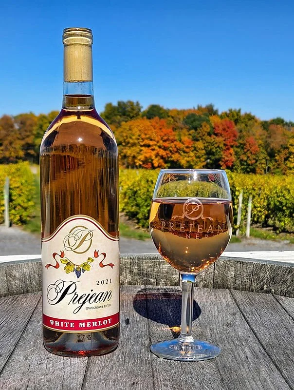 A bottle of 2021 Prejean White Merlot wine and a glass of rosé wine on a wooden surface outdoors with a vineyard and colorful fall trees in the background.