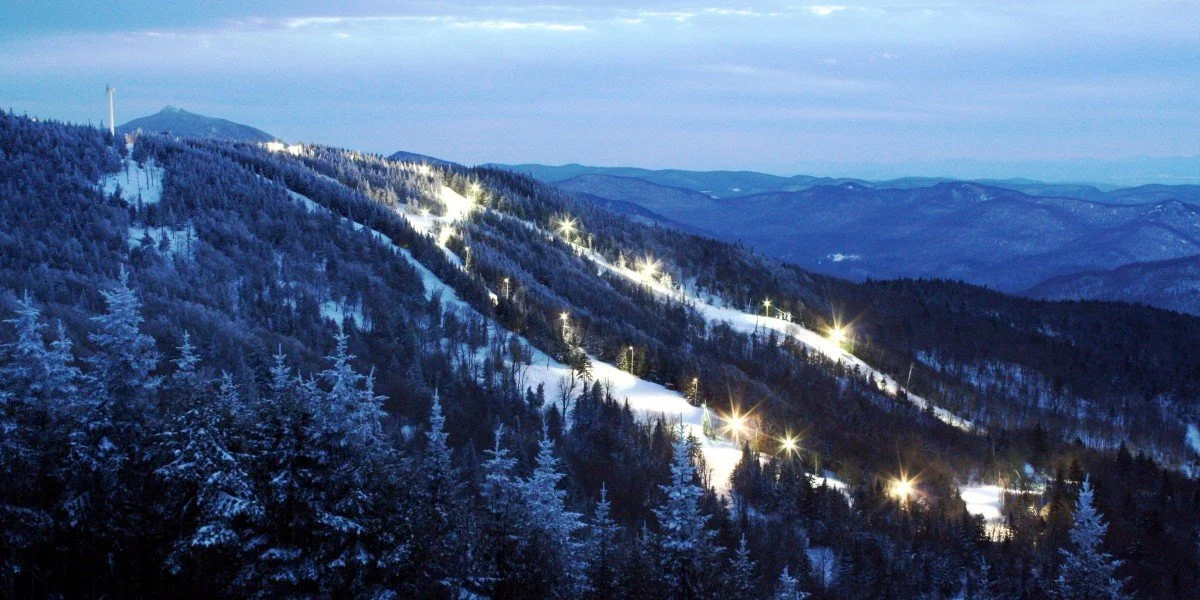 Snow-covered mountain landscape at dusk with ski slopes and ski lifts illuminated by lights.
