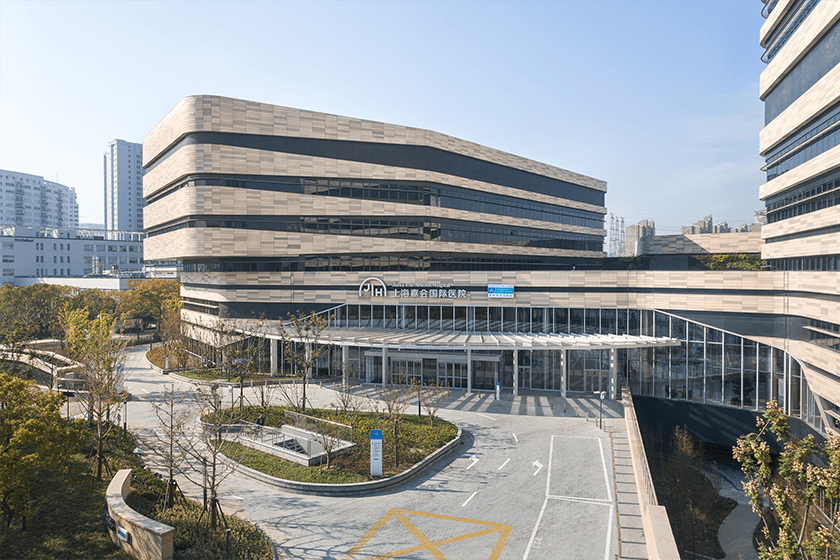 Modern hospital building with glass entrance, surrounded by landscaped trees and parking area, on a sunny day.