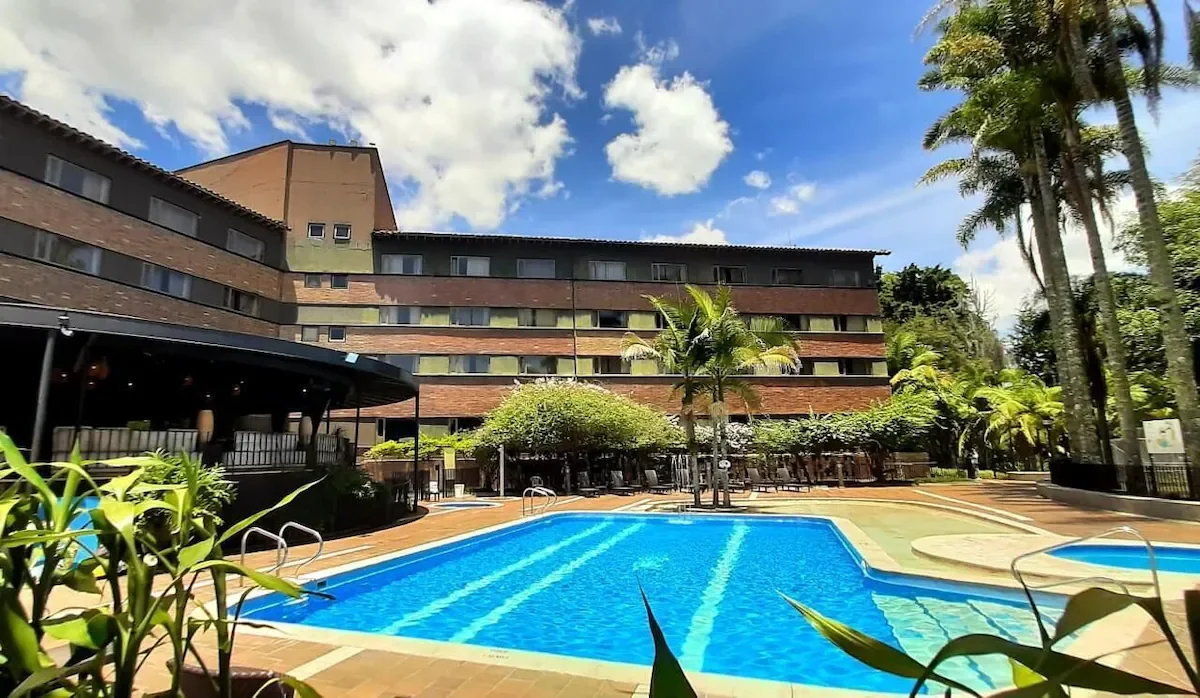 Outdoor swimming pool with blue water, surrounded by a wooden deck, chairs, and lush greenery, in front of a multi-story hotel building under a partly cloudy sky.