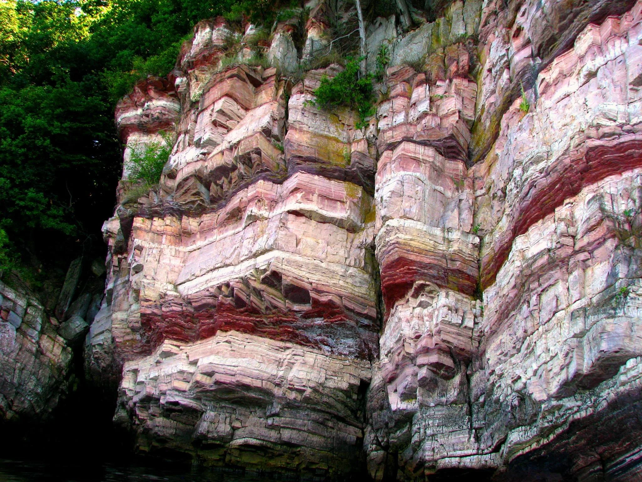 Colorful layered rock formation along a cliff face next to water, with green trees at the top.