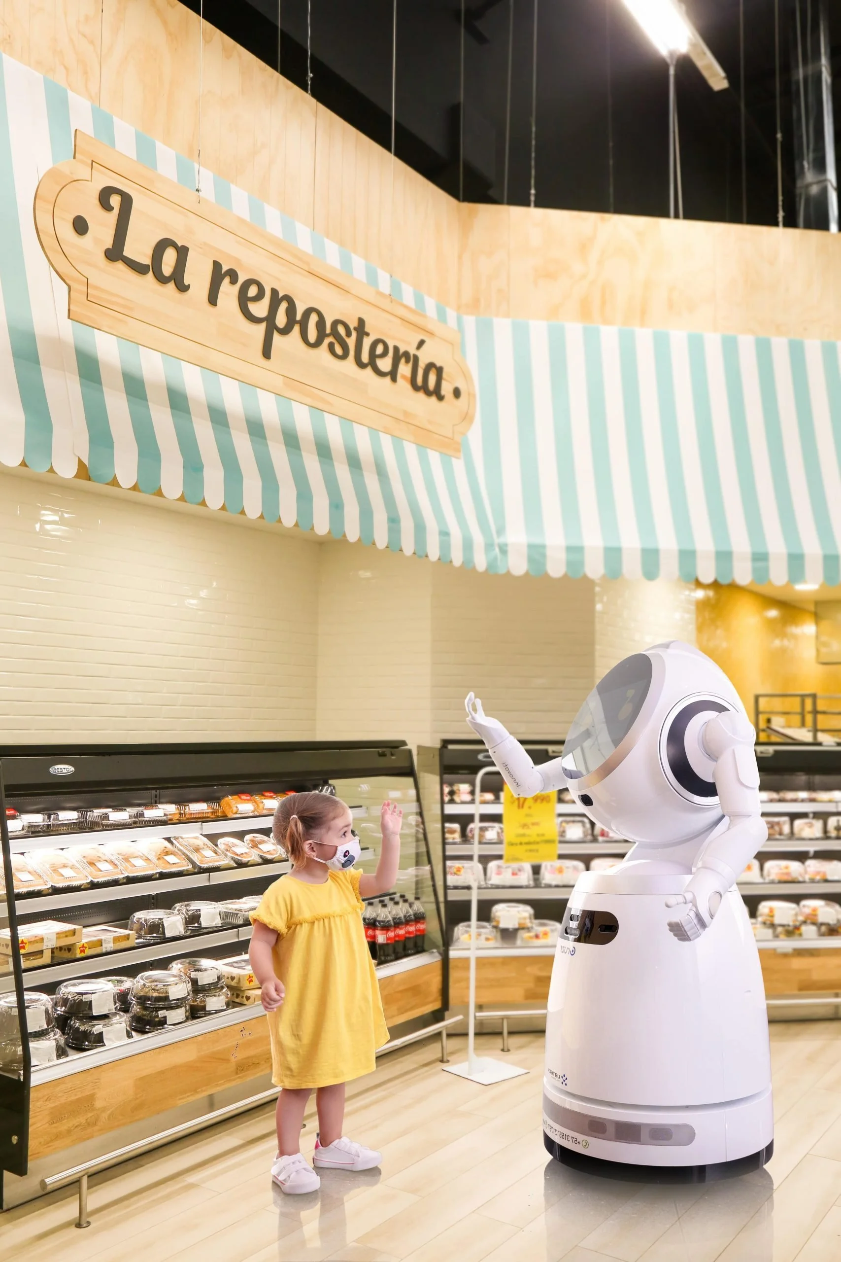 A young girl wearing a yellow dress and a face mask is interacting with a humanoid robot in a grocery store. The girl has her hand raised, possibly waving or reaching out, while the robot is extending its arm towards her. There are shelves with packa