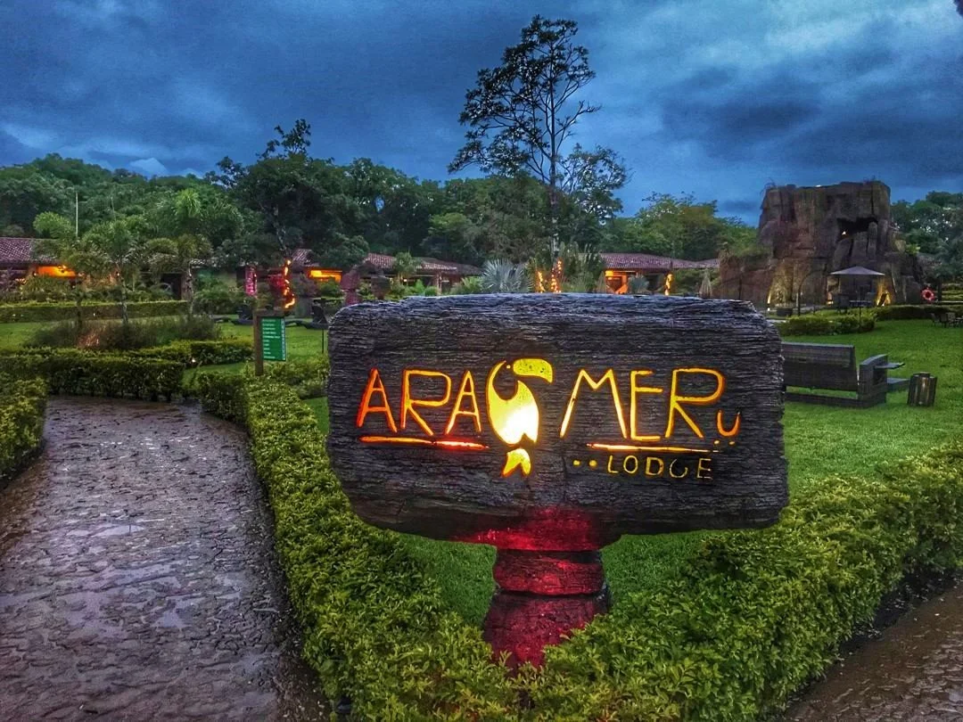 Neon sign for Ara Gumer Lodge on a wooden board at dusk, surrounded by landscaped greenery and a winding stone pathway.