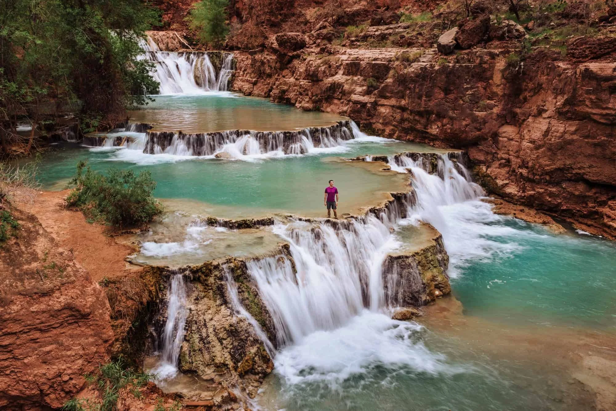 A person standing on a tiered rock formation amidst cascading waterfalls and turquoise water in a canyon surrounded by red cliffs and green vegetation.