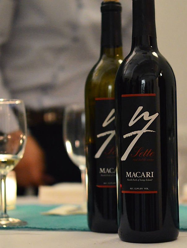 Two bottles of wine, one black labeled 'Sette' and the other green, on a table with wine glasses and a blurred background.