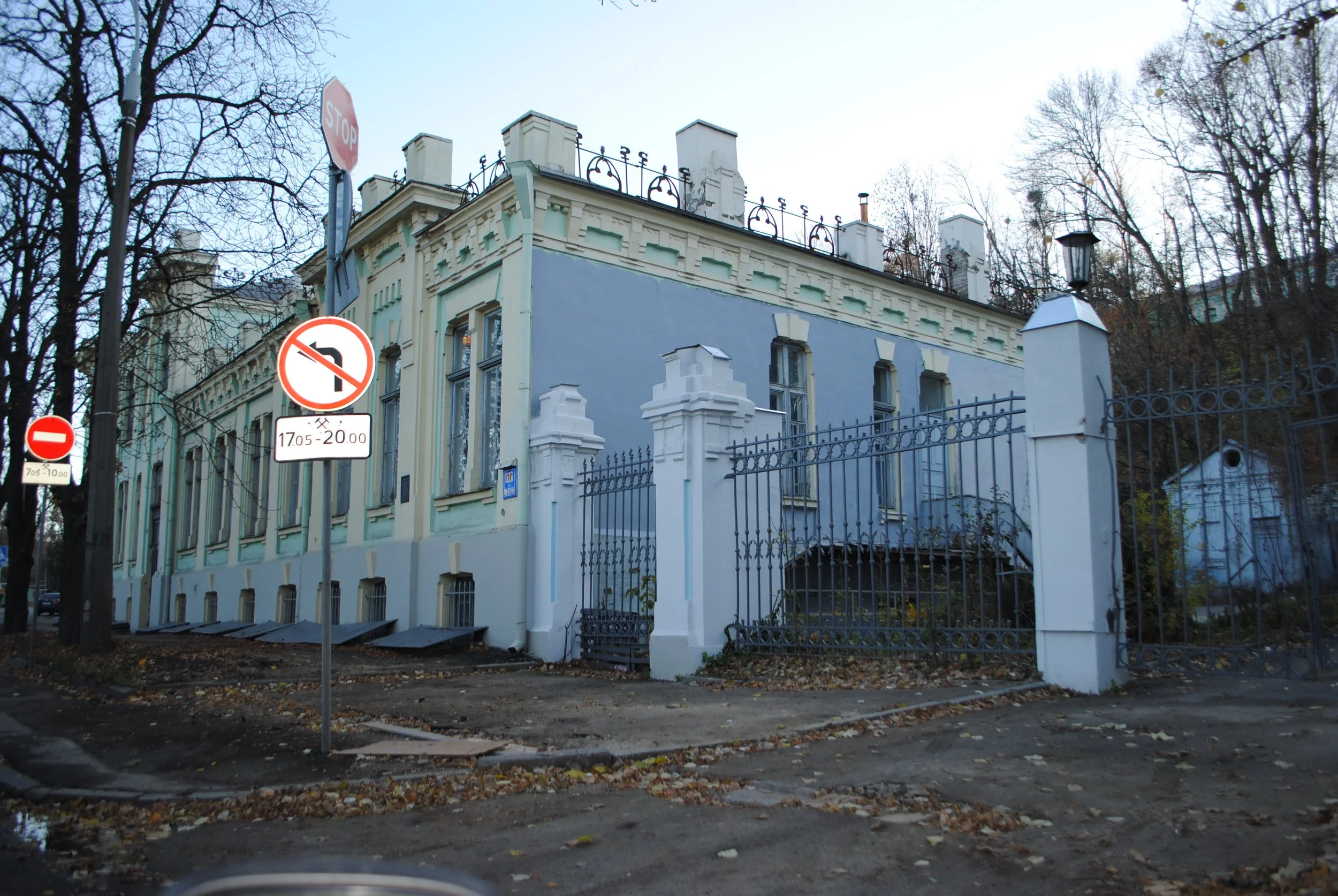 A light blue mansion with decorative white trim, surrounded by a black wrought iron fence. There are leafless trees nearby, and street signs indicating no left turns and restricted parking hours.
