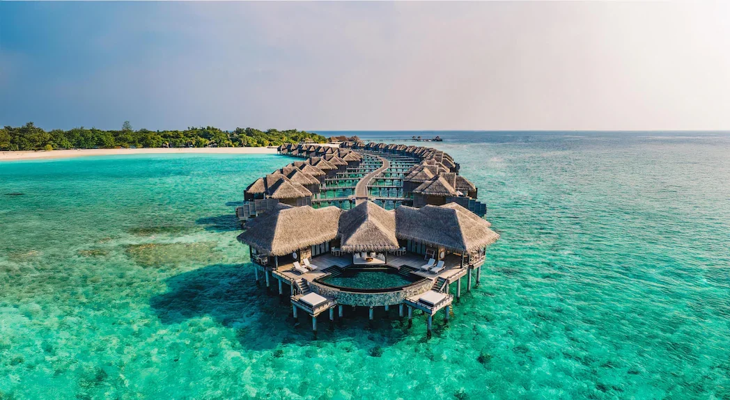 Overwater bungalows with thatched roofs on a clear turquoise ocean, connected by a wooden walkway, with a sandy beach and green trees in the background.