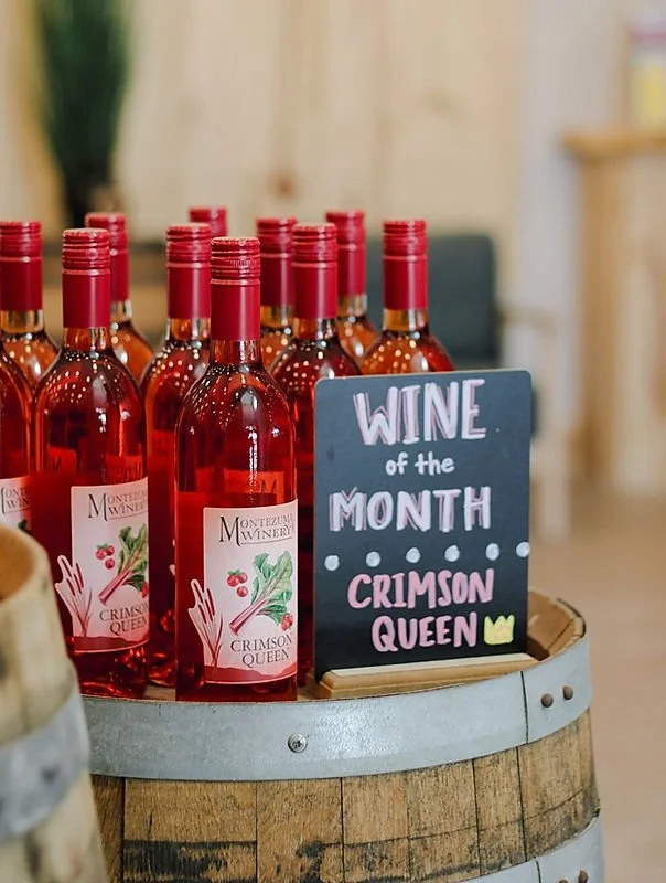 Multiple bottles of pink sparkling wine labeled 'Crimson Queen' on a wine barrel, with a chalkboard sign reading 'Wine of the Month Crimson Queen'.
