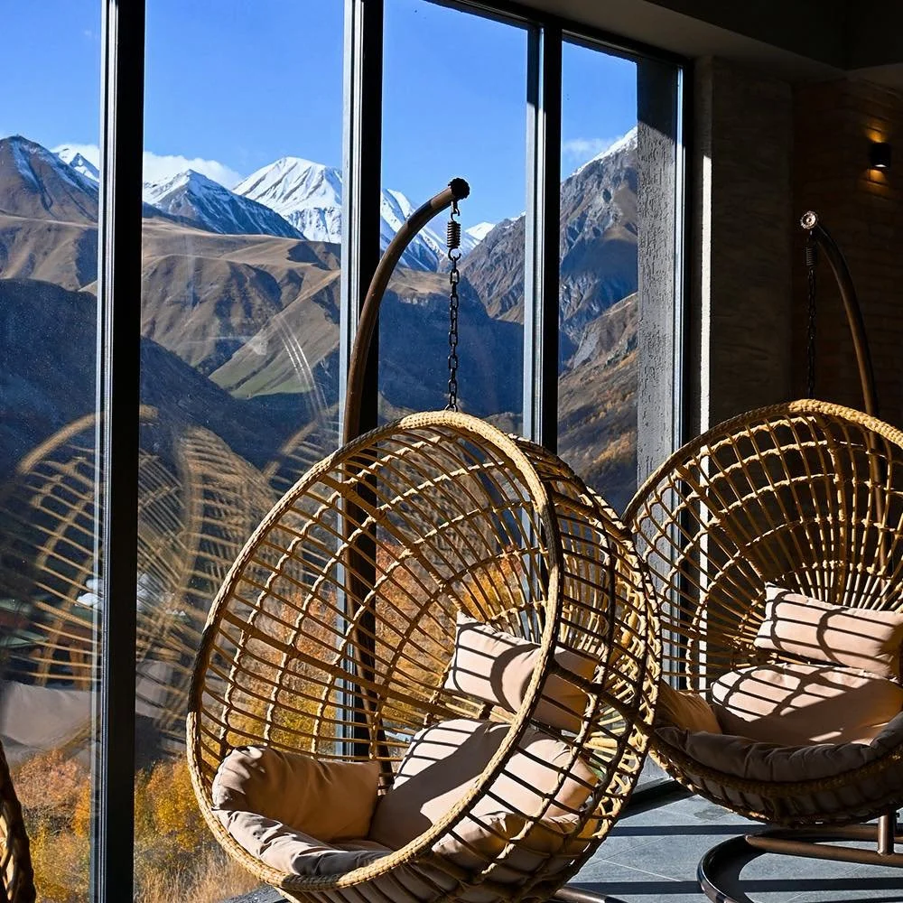Indoor seating area with hanging rattan chairs and beige cushions, large glass windows showing mountain landscape with snow-capped peaks and clear blue sky.