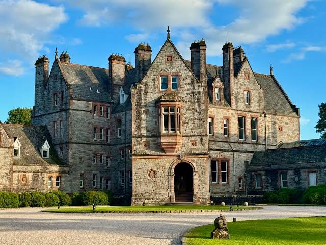 A historic stone castle with multiple turrets and pointed rooftops, set against a partly cloudy blue sky, surrounded by manicured lawns.