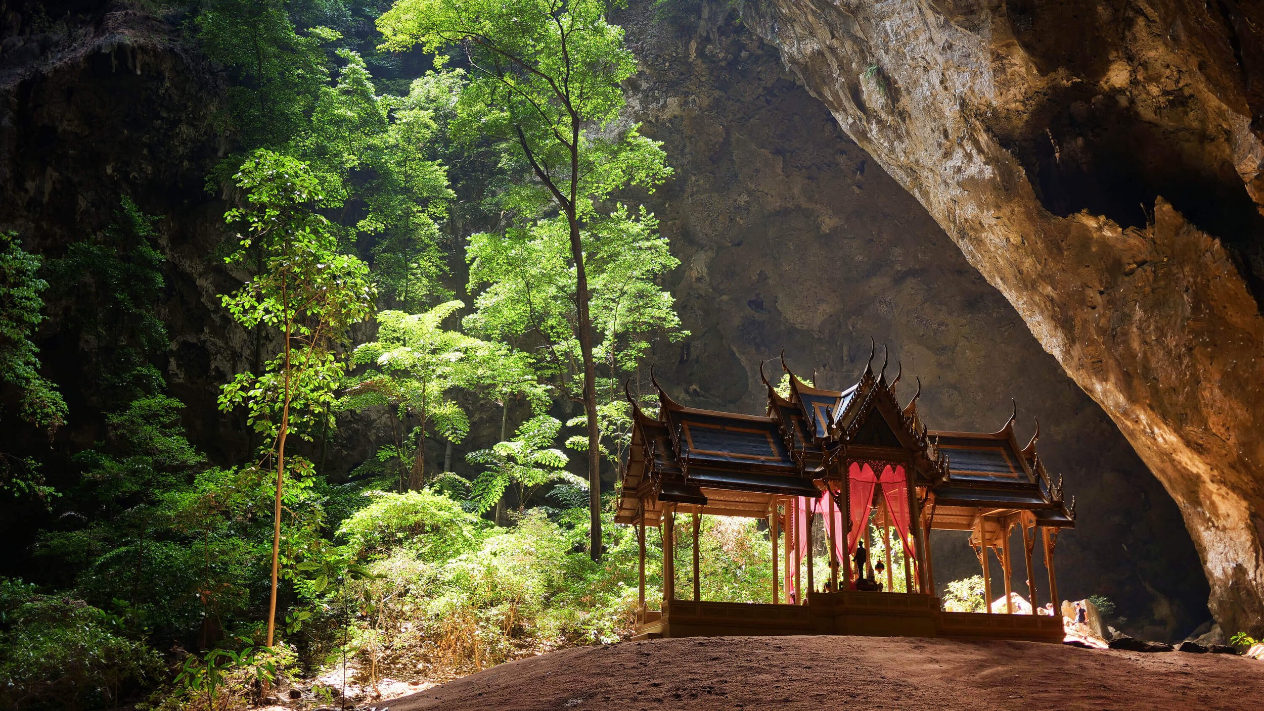 A temple with traditional Asian architecture inside a large cave with lush green trees and rock formations visible.
