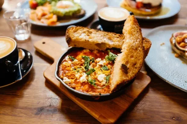 Baked kamado pizza with tomato sauce, cheese, and herbs, served with toasted bread slices on a wooden board, surrounded by plates of food, cups of coffee, and a small bowl of nuts.