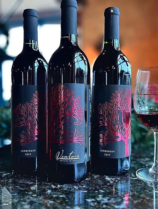 Three bottles of red wine with dark labels featuring pink tree illustrations, labeled "Lemberger 2019" from Ventosa Estate Winery, and a glass of red wine on a reflective surface.