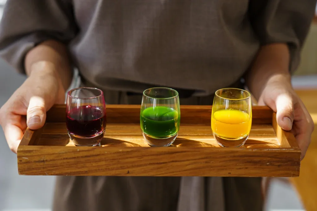 Person holding a wooden tray with three shot glasses filled with red, green, and yellow liquids.