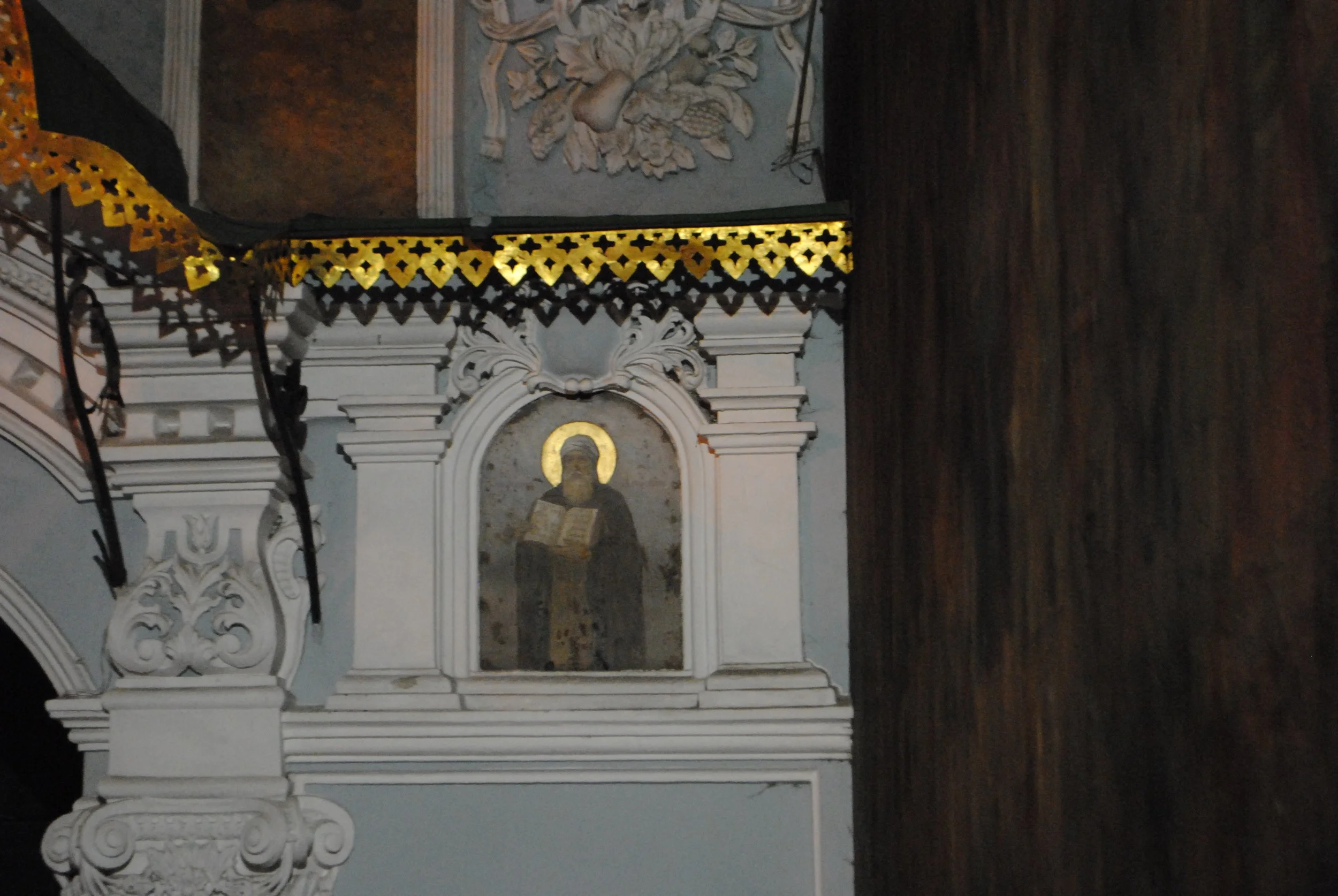 An ornate altar with white decorative carvings, gold and black patterned trim, and a religious icon of a saint with a halo holding a book. The altar has intricate details and contrasts with a dark wooden background.
