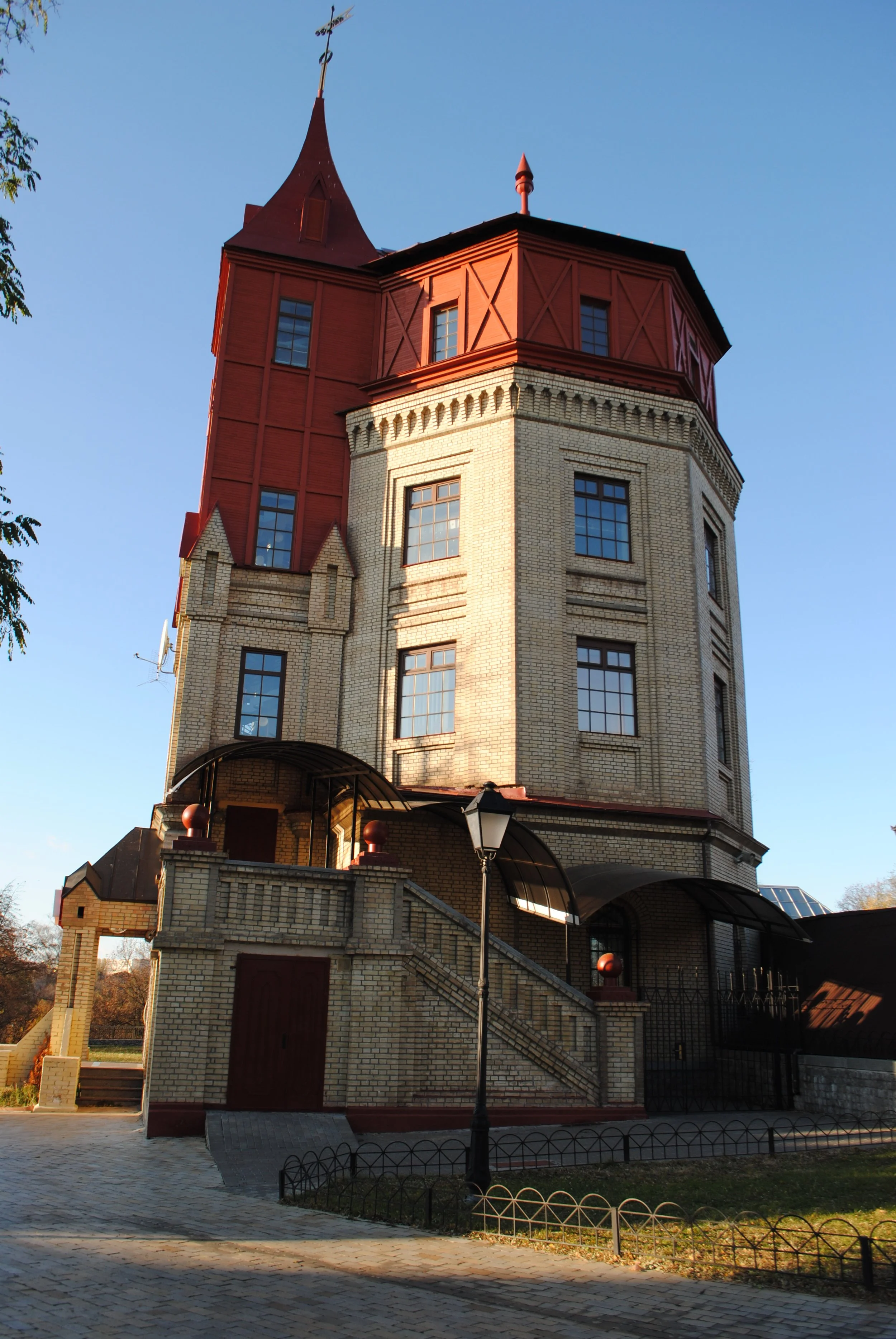 A multi-story building with a beige brick facade and a prominent red roof, featuring a tower-like structure with several windows, a small balcony, and decorative architectural details. The scene is set outdoors with a clear blue sky, a lamp post, a p