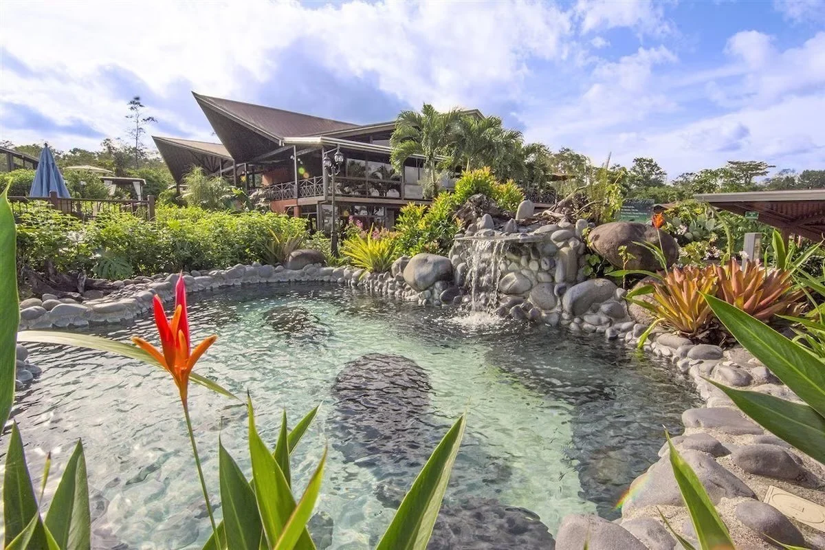 A tropical outdoor scene featuring a stone-lined swimming pool with a small waterfall, surrounded by lush green plants and flowers, with a wooden building in the background and a partly cloudy sky overhead.