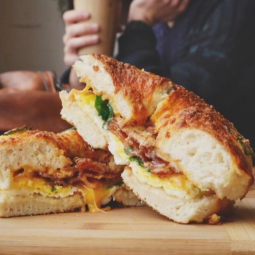 Close-up of a cut sandwich with melted cheese, eggs, bacon, and green vegetables on a wooden cutting board, with a person holding a drink in the background.