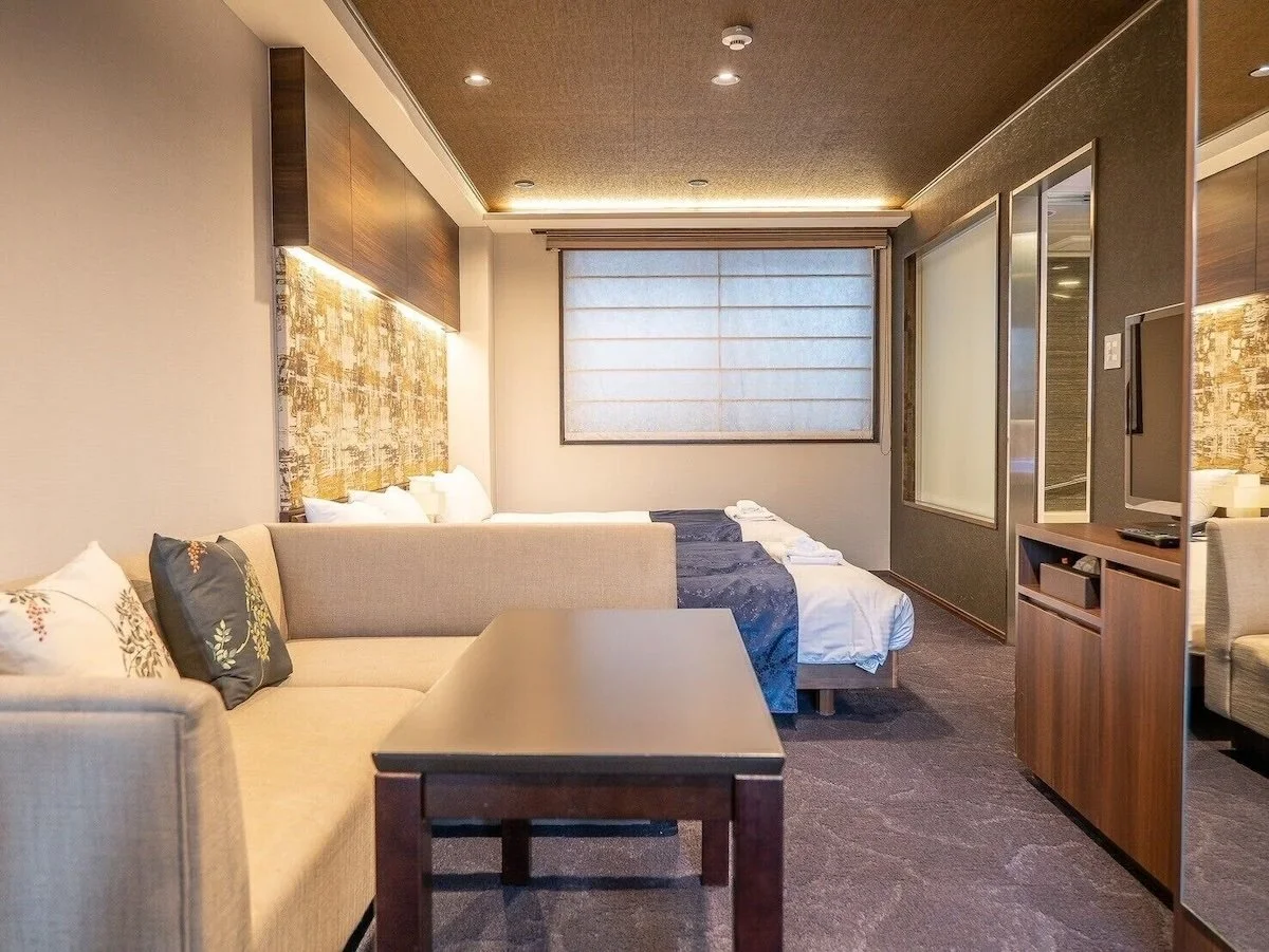 Hotel room with bed, sofa, coffee table, television, and window with blinds.
