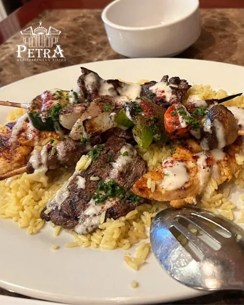 Plate of grilled chicken and beef skewers with rice, topped with creamy white sauce, on a white plate with a fork. In the background, a small white bowl and a logo of Petra Mediterranean Bistro.