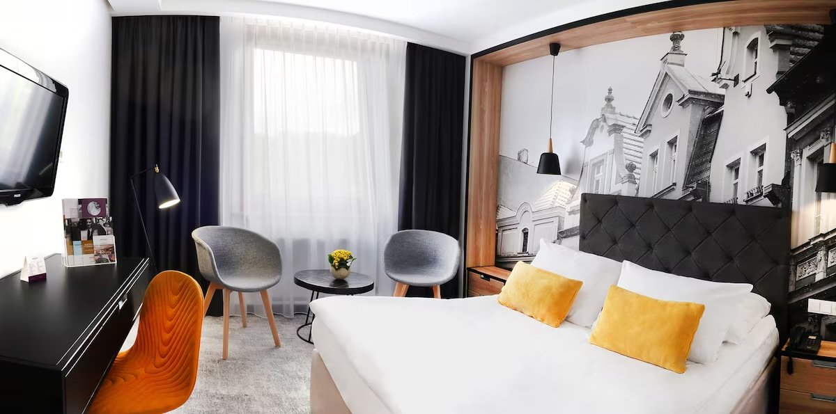 Hotel room with bed, black upholstered headboard, black and white mural of city buildings, two gray chairs, black round table with yellow flowers, black desk with an orange chair, window with sheer white and black curtains, wall-mounted TV.