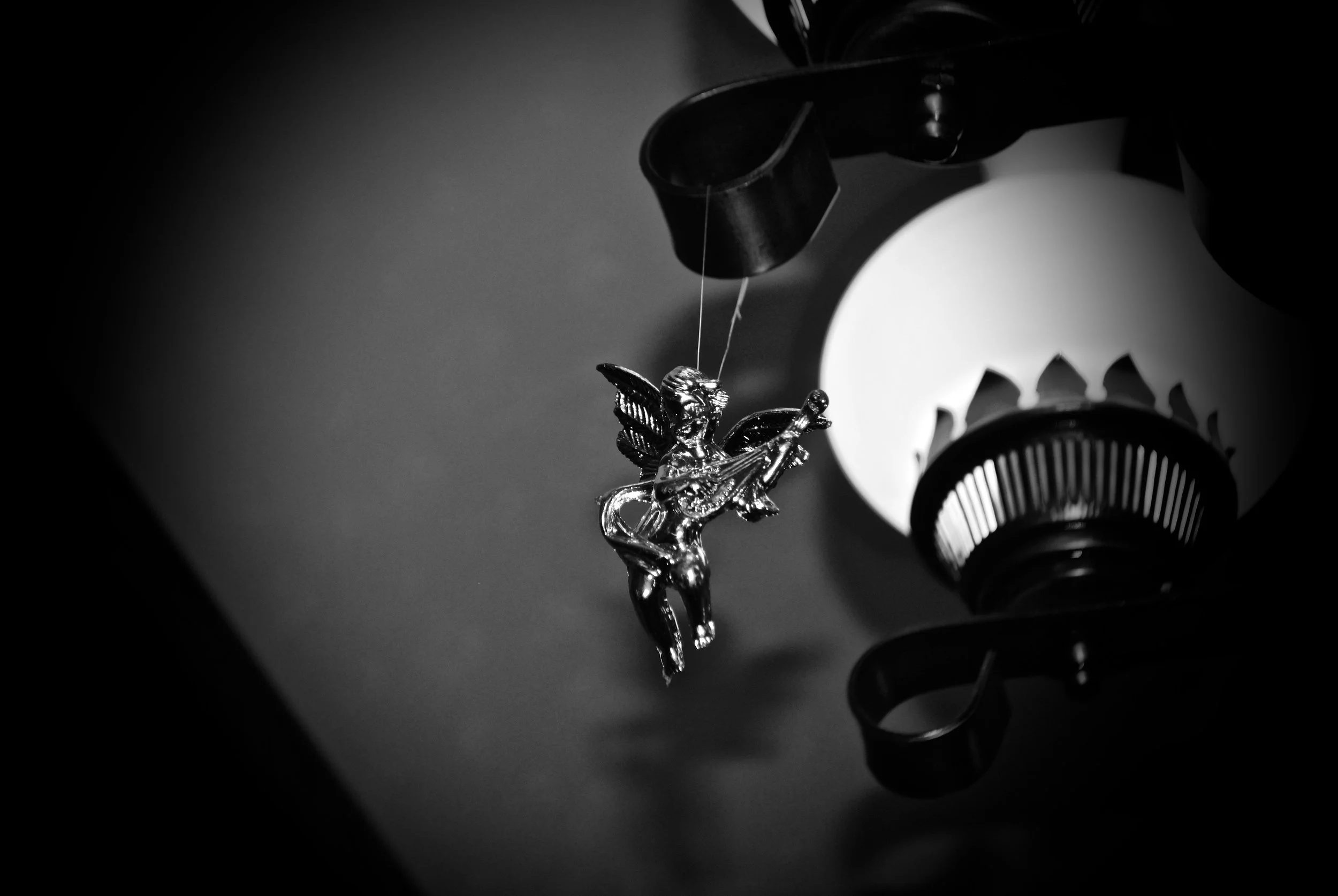 Black and white close-up of a vintage cigarette lighter with a cherub playing a musical instrument hanging from it and a light fixture in the background.