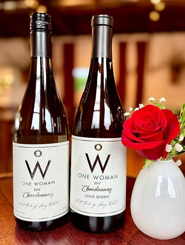 Two bottles of wine labeled 'One Woman,' with a single red rose in a white vase next to them.