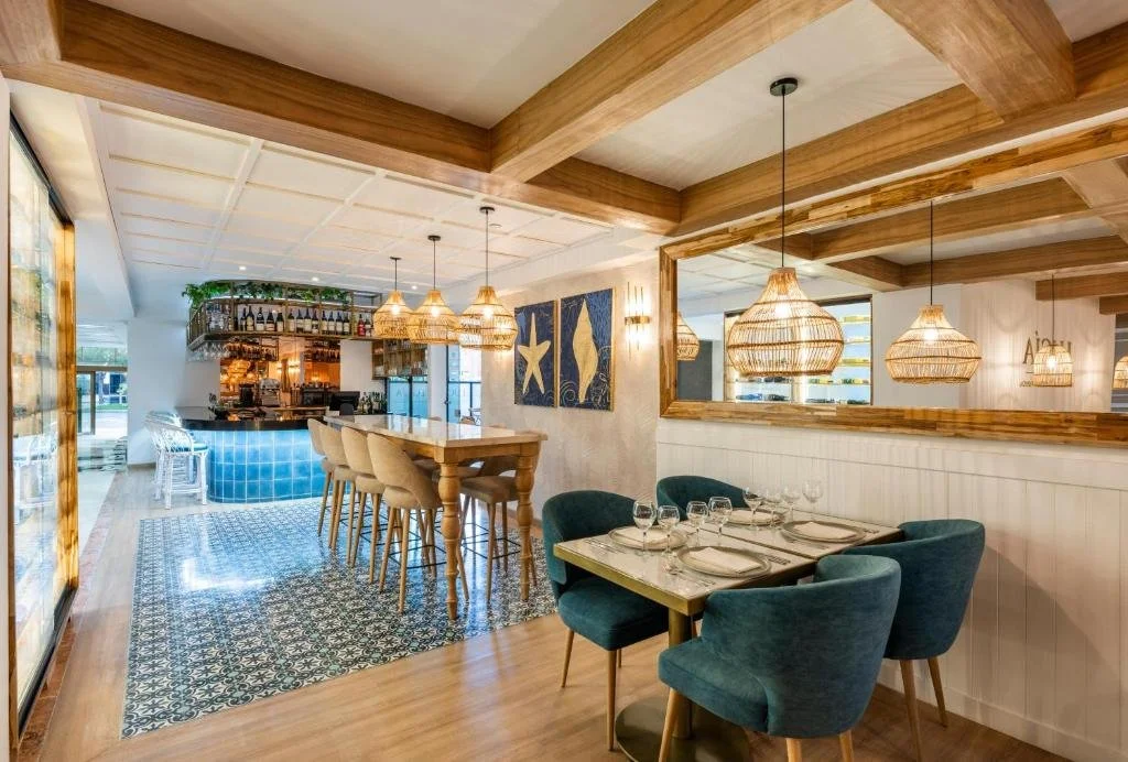 Elegant restaurant interior with a bar area and a dining table set for four, featuring teal upholstered chairs, wooden accents, wicker pendant lights, and nautical artwork.