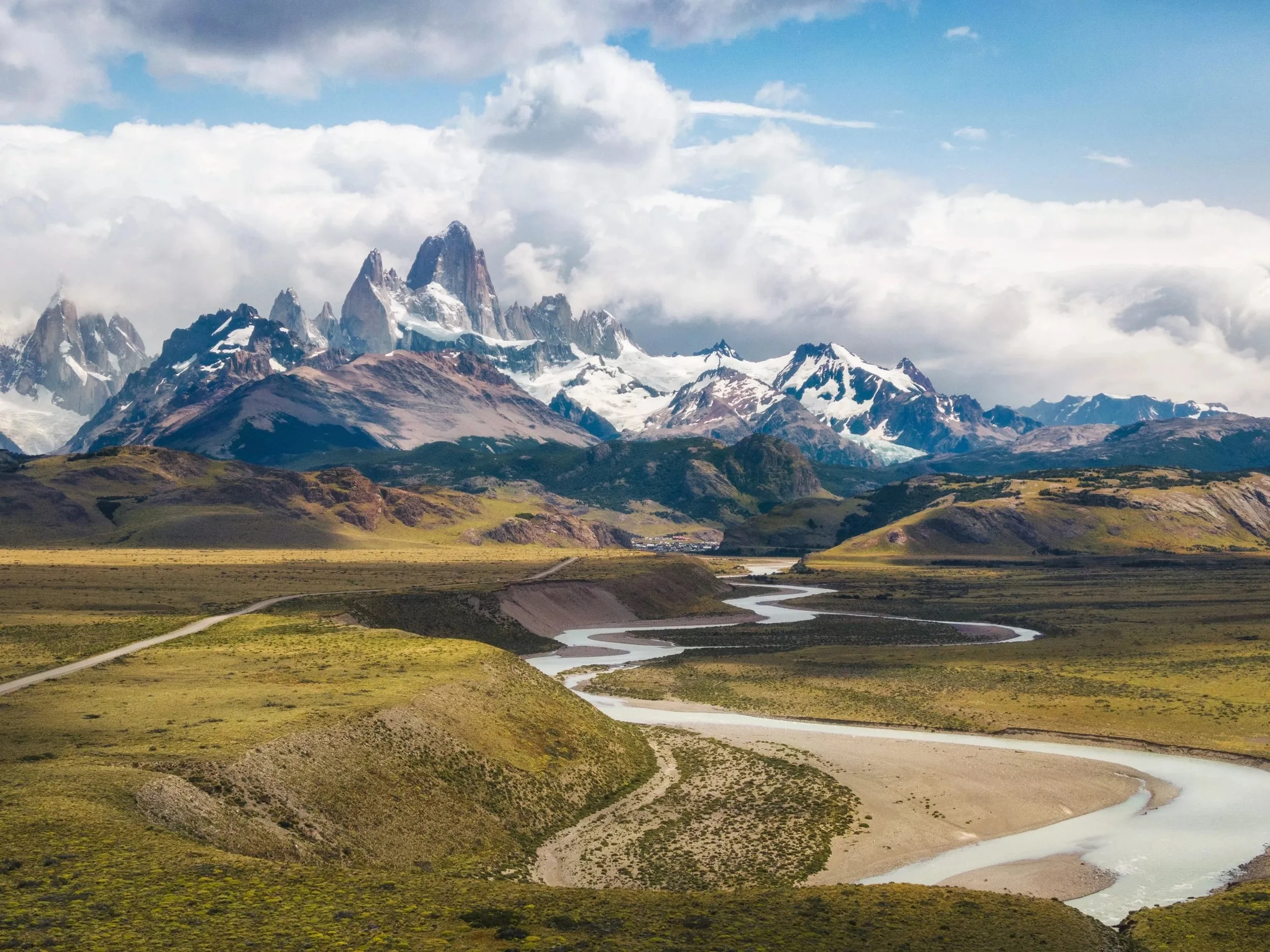 Snow-capped mountains with rocky peaks in the background, grassy hills in the foreground, and a winding river flowing through the valley below under a partly cloudy sky.
