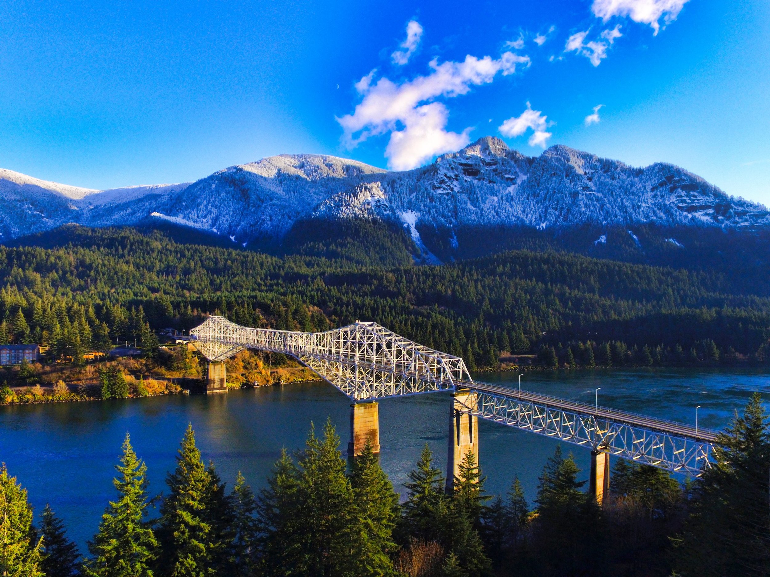 A mountain landscape with a snow-capped mountain range, a forest, a river, and a bridge over the river.