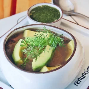 Bowl of cold soup garnished with avocado slices and fresh herbs, with a small cup of green sauce or salsa in the background.