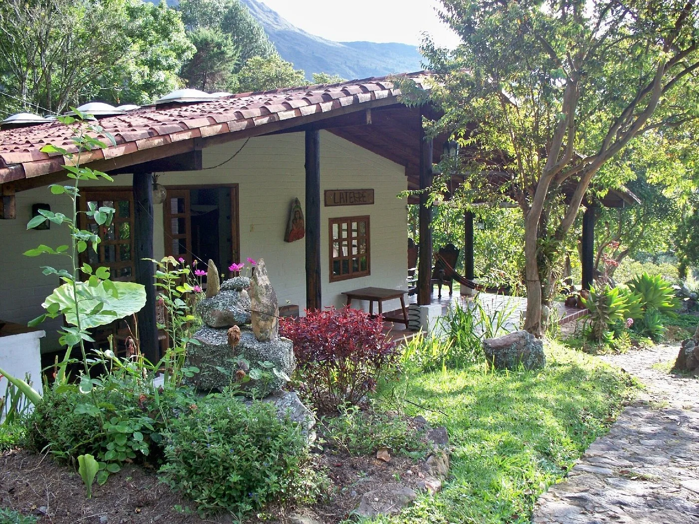 A small house with a tiled roof, surrounded by greenery and trees, with a stone pathway and a garden in the foreground.