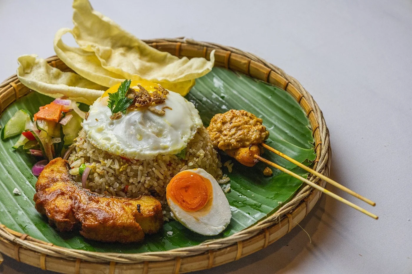Indonesian-style fried rice dish served on a banana leaf in a wicker basket, garnished with a fried egg, half a boiled egg, fried chicken, satay skewers, crackers, and fresh vegetable salad.