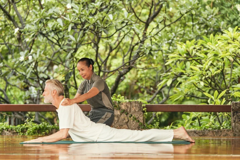 A woman assisting a man in a yoga pose on a mat outdoors with lush green trees in the background.