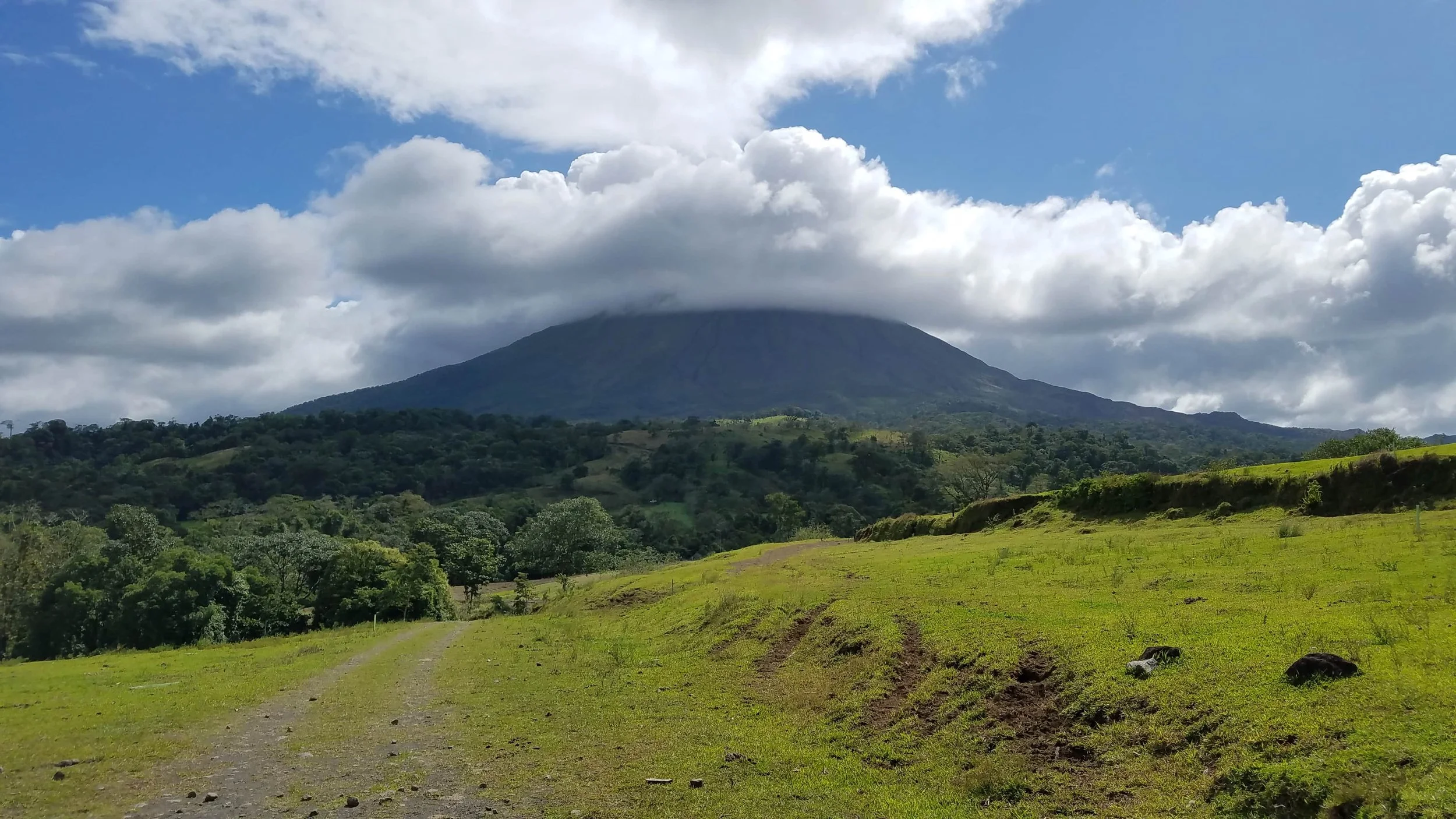 A lush green landscape with a dirt pathway in the foreground, rolling hills covered in trees in the midground, and a large volcano with a cloud on top in the background under a partly cloudy sky.