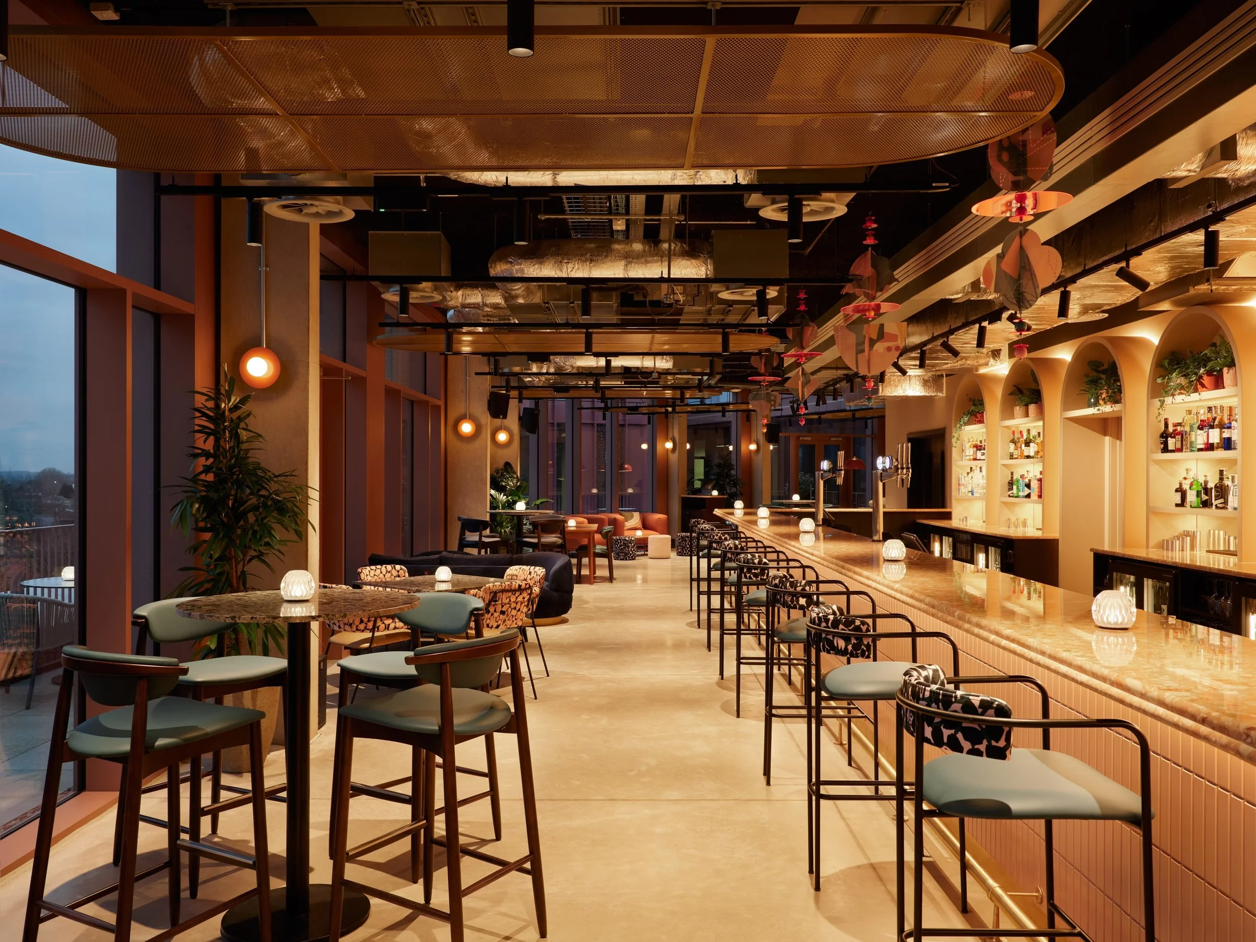 Interior of a modern bar or restaurant with high tables, bar stools, a bar counter, and decorative lighting fixtures on the ceiling.