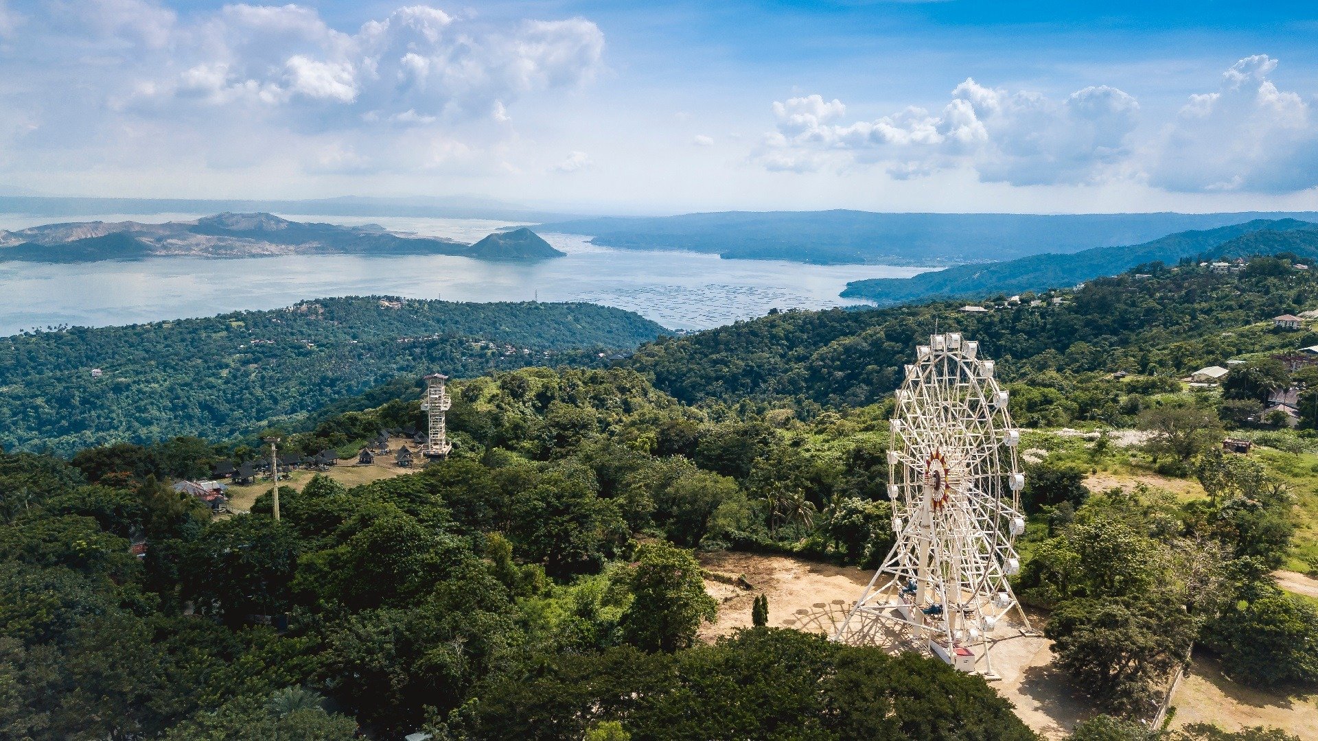 A lush green landscape with rolling hills, a large white Ferris wheel, and a charming small town. A body of water with an island and distant mountains is visible in the background, under a partly cloudy sky.