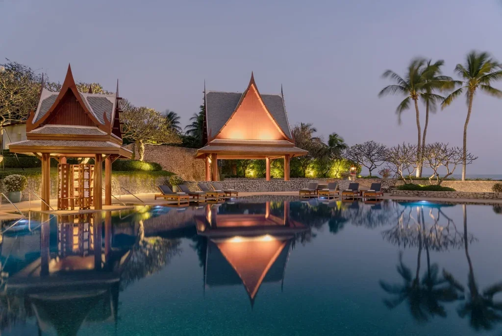 A tranquil swimming pool with a thatched-roof pavilion and lounge chairs, surrounded by palm trees and tropical plants at dusk.