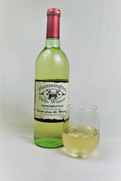 A green bottle of Hummingbird Hills Winery wine with a pink top, next to a glass filled with white wine.