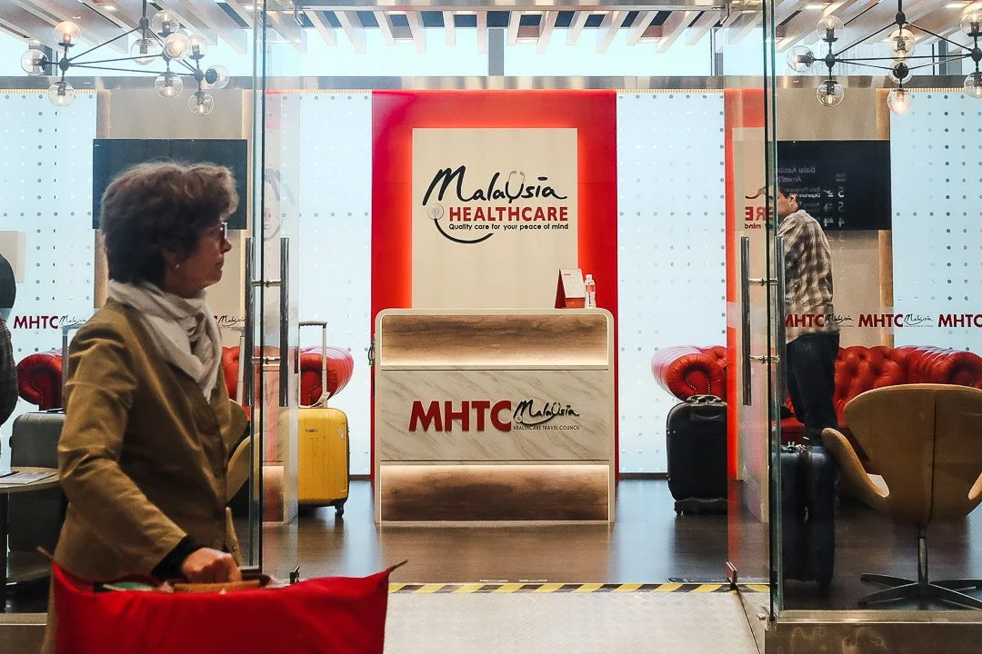Interior of a Malaysian HealthcareTravel Council office with a reception desk, red and white branding, and seating area, seen through glass doors with people walking inside.
