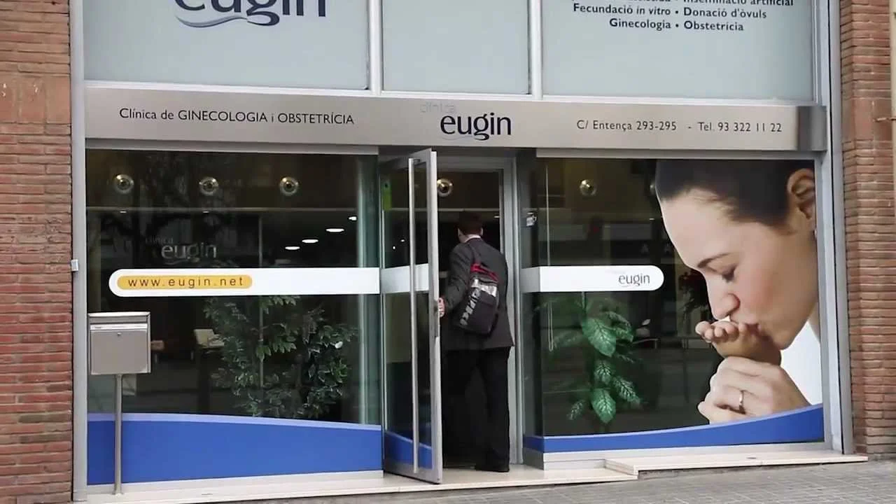 Exterior of a gynecology and obstetrics clinic with large glass windows, a man entering the building, and a large promotional image of a woman praying on the right window.