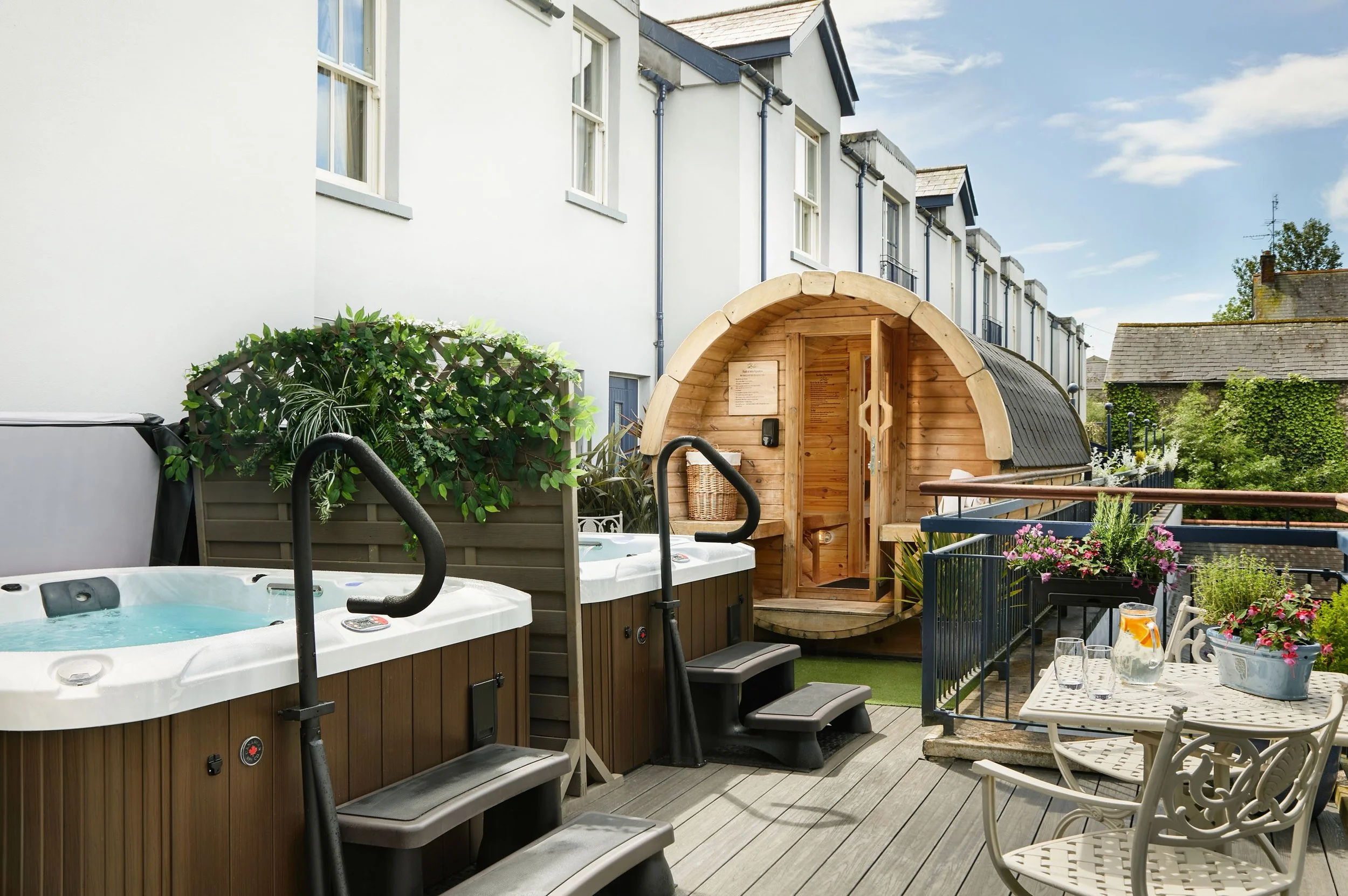 A backyard patio with hot tubs, a wooden sauna, dining table with drinks, and lush greenery outside a white residential building.