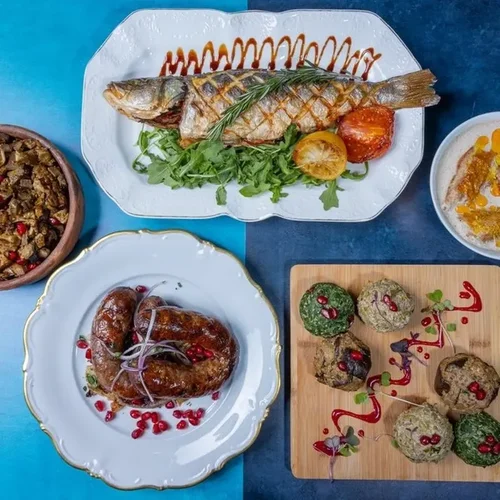 A variety of dishes including grilled fish with lemon and greens, roasted chicken with pomegranate seeds, a bowl of cooked meat or vegetables, and stuffed grape leaves with garnishes, served on decorative plates and a wooden board.